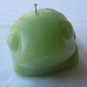 May include: A green frog-shaped candle with a white wick. The candle has a smooth, rounded surface and two large, round eyes.