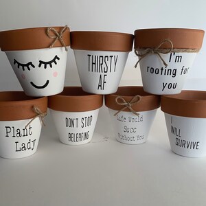 May include: Set of eight white terracotta pots with various plant-themed sayings. The sayings include "Plant Lady", "Don't Stop Beleafing", "Life Would Succ Without You", "I Will Survive", "Thirsty AF", "I'm Rooting For You", and two pots with a smiley face and eyelashes.