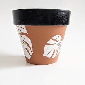 Puede incluir: Una maceta de terracota con un borde negro y hojas tropicales pintadas de blanco.