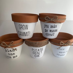 May include: Five white terracotta flower pots with brown rims and twine accents. The pots are decorated with black text, including "Grow Dammit!", "You make me wanna sprout!", "Plant one on me", "Please don't die", and "Once upon a thyme".