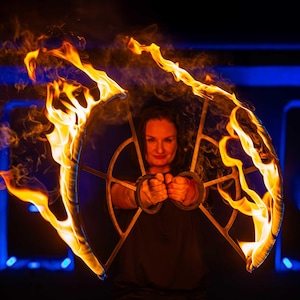 Peut inclure: Une femme tient un accessoire circulaire en flammes. L'accessoire a un centre bleu foncé avec un cadre doré. Des flammes orange et jaunes vives entourent l'accessoire, créant un effet visuel saisissant sur un fond sombre.