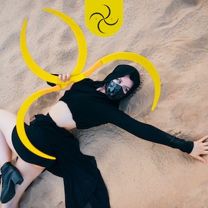 Pode incluir: Uma pessoa em um traje preto com capuz e uma máscara facial geométrica está deitada na areia, segurando dois grandes objetos amarelos em forma de crescente. Um emblema amarelo com um design em espiral está acima. A pessoa está usando botas pretas.