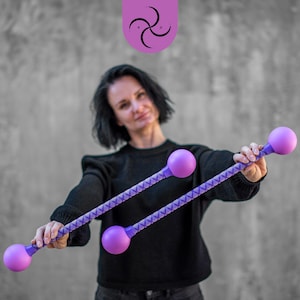 Könnte beinhalten: Eine Frau hält zwei lila Jonglierstöcke mit lila Kugeln an den Enden. Die Stöcke haben ein lila-weißes Muster.