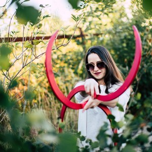 以下が含まれることがあります： サングラスと白いシャツを着た女性が、庭で大きな赤い金属製の輪2つを持ってポーズをとっている。