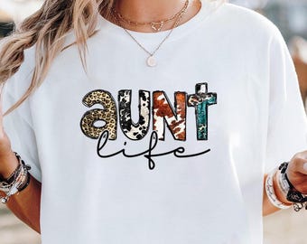 May include: White t-shirt with the words "AUNT life" in a mix of animal print and turquoise lettering. The word "AUNT" is in a bold, decorative font, while "life" is in a cursive script. The shirt is a casual style.