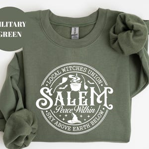 May include: A military green crewneck sweatshirt featuring a white circular graphic. The text reads Local Witches Union, Salem, Peace Within, and Sky Above Earth Below, accompanied by illustrations of a cauldron, broomsticks, bats, and a hat.