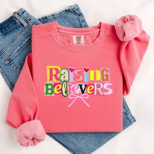 Peut inclure: Un sweat-shirt corail avec le texte "Raising Believers" dans un style de lettres en blocs color&eacute;s. Le sweat-shirt a un col rond et des manches &agrave; revers. Une paire de jeans bleus est visible en arri&egrave;re-plan.