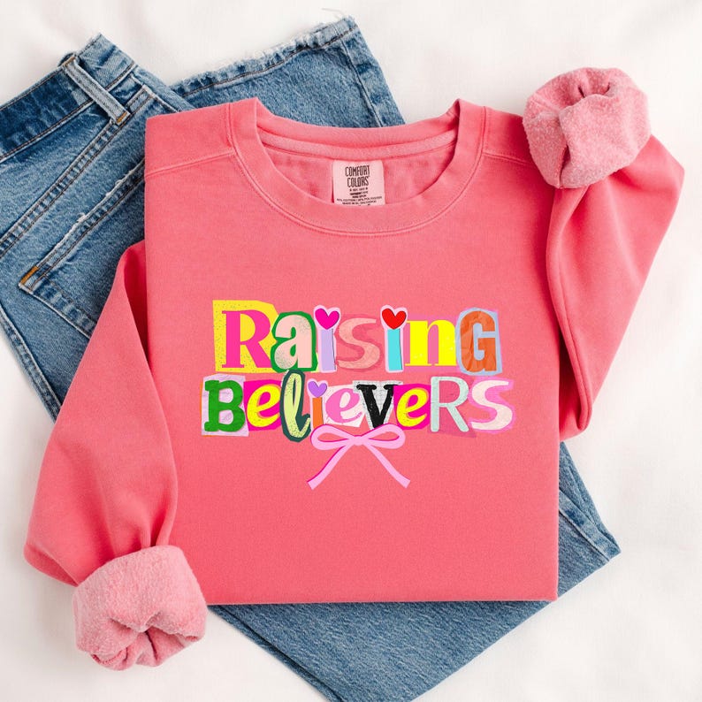 Peut inclure: Un sweat-shirt corail avec le texte "Raising Believers" dans un style de lettres en blocs color&eacute;s. Le sweat-shirt a un col rond et des manches &agrave; revers. Une paire de jeans bleus est visible en arri&egrave;re-plan.