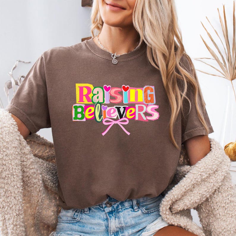 Raising Believers graphic shirt featuring playful layered typography with mixed font styles and decorative heart accents; the design displays the phrase Raising Believers in bold stacked lettering with textured details and a bow graphic centered beneath the text; the layout is balanced and easy to read with a cheerful faith inspired aesthetic; the artwork is centered on the shirt creating a clean modern graphic suitable for everyday wear