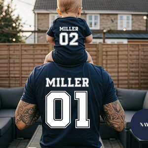 May include: Navy blue matching shirts with the name "MILLER" and numbers "01" and "02" in white. The "02" shirt is a baby onesie. The "01" shirt is an adult t-shirt. The shirts are worn by a parent and child.