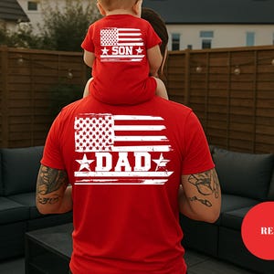 Puede incluir: Camisetas rojas con un dise&ntilde;o de bandera americana desgastada. La camiseta m&aacute;s grande dice "DAD" y la m&aacute;s peque&ntilde;a dice "SON". Las camisetas las llevan un padre y un hijo, con el ni&ntilde;o sentado sobre los hombros del padre. Las camisetas son un conjunto a juego.
