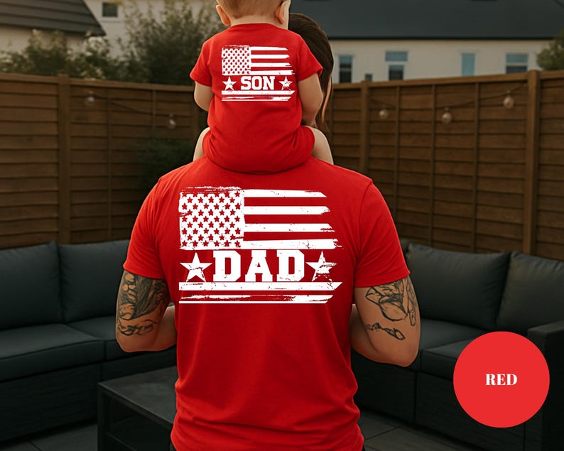 Puede incluir: Camisetas rojas con un dise&ntilde;o de bandera americana desgastada. La camiseta m&aacute;s grande dice "DAD" y la m&aacute;s peque&ntilde;a dice "SON". Las camisetas las llevan un padre y un hijo, con el ni&ntilde;o sentado sobre los hombros del padre. Las camisetas son un conjunto a juego.