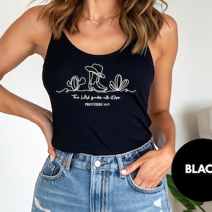 May include: Black spaghetti strap tank top with a white graphic of a cowboy hat, boots, and cactus. The text "The Lord guides not stops" and "PROVERBS 16:9" are below the graphic. The model is wearing blue denim jeans.