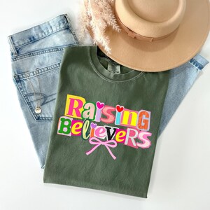 Raising Believers graphic shirt featuring playful layered typography with mixed font styles and decorative heart accents; the design displays the phrase Raising Believers in bold stacked lettering with textured details and a bow graphic centered beneath the text; the layout is balanced and easy to read with a cheerful faith inspired aesthetic; the artwork is centered on the shirt creating a clean modern graphic suitable for everyday wear