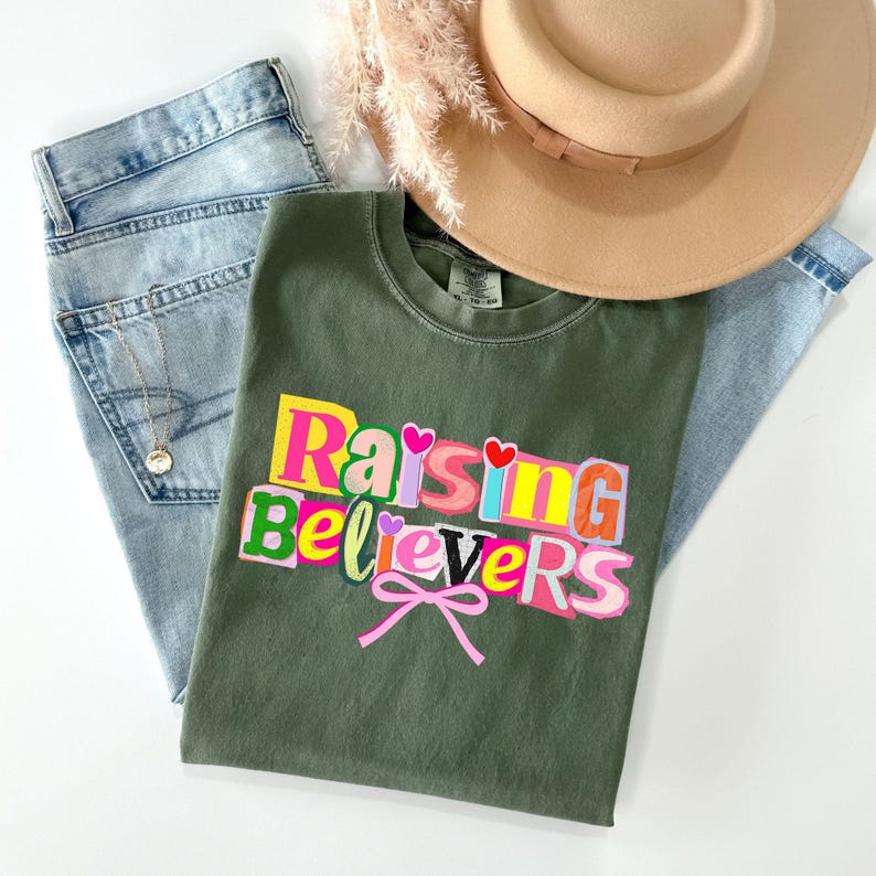 Raising Believers graphic shirt featuring playful layered typography with mixed font styles and decorative heart accents; the design displays the phrase Raising Believers in bold stacked lettering with textured details and a bow graphic centered beneath the text; the layout is balanced and easy to read with a cheerful faith inspired aesthetic; the artwork is centered on the shirt creating a clean modern graphic suitable for everyday wear