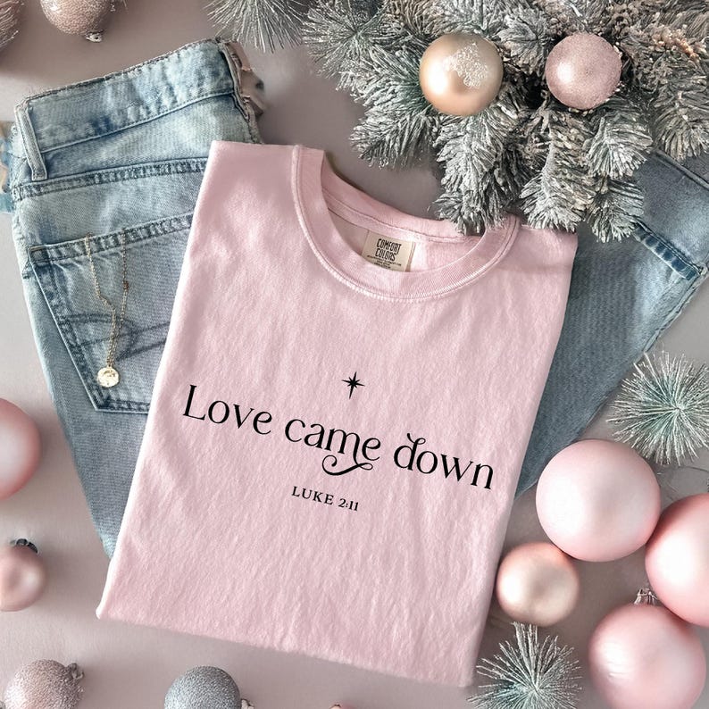May include: A pale pink t-shirt with the words "Love came down" and "Luke 2:11" printed in black. The shirt is placed on blue denim jeans, with Christmas baubles and decorative branches.
