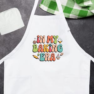 May include: White apron with two front pockets and the text "IN MY BAKING ERA" in colorful retro font. A white chef's hat is in the upper left corner. A green and white checkered cloth is in the upper right corner.