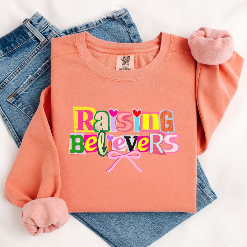 Peut inclure: Un sweat-shirt corail avec le texte "Raising Believers" en lettres color&eacute;es de style d&eacute;coup&eacute;. Le sweat-shirt a un col rond et des manches longues avec des poignets roses. Le sweat-shirt est pos&eacute; sur un jean bleu.
