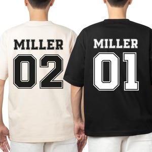 May include: Two short-sleeved t-shirts, one cream and one black, with the name "MILLER" and the numbers "02" and "01" printed on the back in a collegiate style. The numbers are outlined in a contrasting color.
