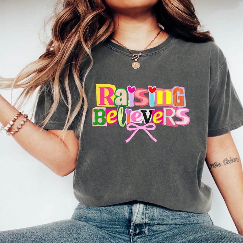 Raising Believers graphic shirt featuring playful layered typography with mixed font styles and decorative heart accents; the design displays the phrase Raising Believers in bold stacked lettering with textured details and a bow graphic centered beneath the text; the layout is balanced and easy to read with a cheerful faith inspired aesthetic; the artwork is centered on the shirt creating a clean modern graphic suitable for everyday wear
