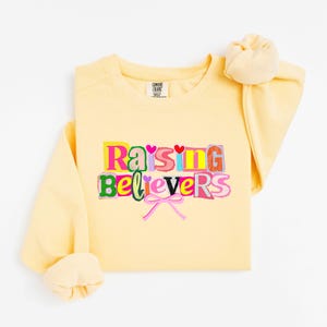 Raising Believers graphic shirt featuring playful layered typography with mixed font styles and decorative heart accents; the design displays the phrase Raising Believers in bold stacked lettering with textured details and a bow graphic centered beneath the text; the layout is balanced and easy to read with a cheerful faith inspired aesthetic; the artwork is centered on the shirt creating a clean modern graphic suitable for everyday wear