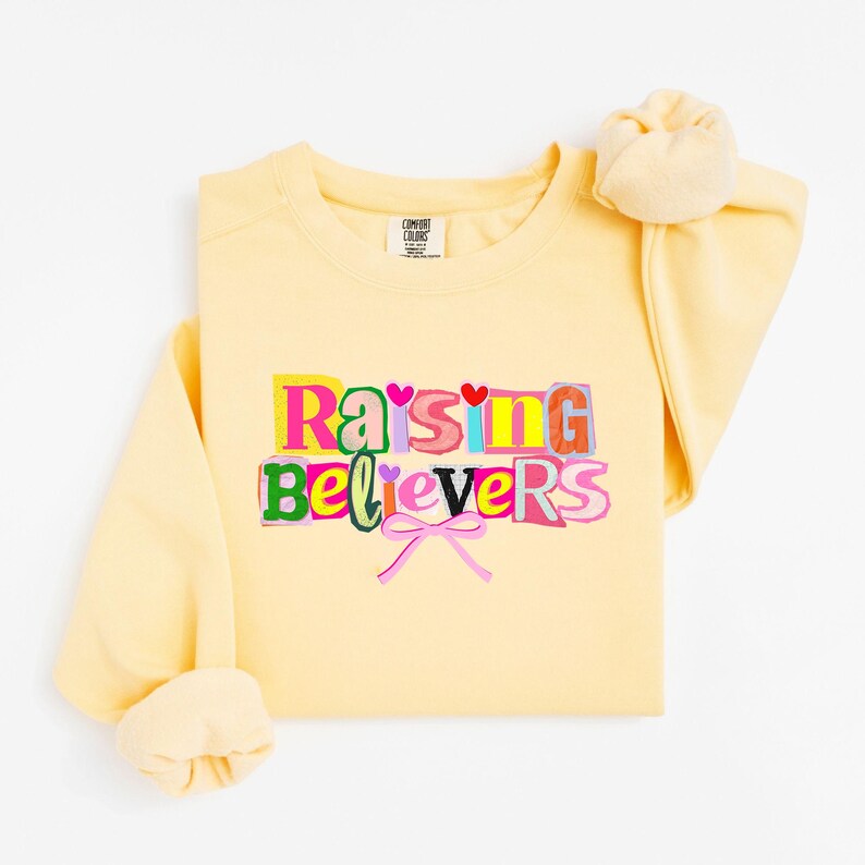 Raising Believers graphic shirt featuring playful layered typography with mixed font styles and decorative heart accents; the design displays the phrase Raising Believers in bold stacked lettering with textured details and a bow graphic centered beneath the text; the layout is balanced and easy to read with a cheerful faith inspired aesthetic; the artwork is centered on the shirt creating a clean modern graphic suitable for everyday wear