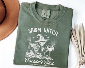 May include: An olive green t-shirt featuring a white graphic print of cocktails, a witch hat, a skull, and pumpkins. The text reads SALEM WITCH COCKTAIL CLUB and SALEM SPIRITS, PICK YOUR POISON. The shirt has a Comfort Colors brand label in the neck.