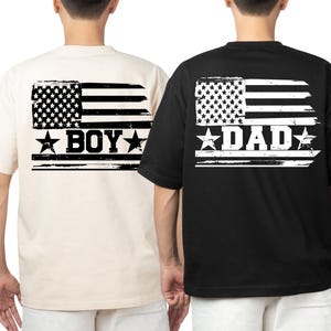 Puede incluir: Dos camisetas, una crema y otra negra, presentan un dise&ntilde;o de bandera americana desgastada en la espalda. La camiseta crema dice "BOY" y la negra dice "DAD" en letras blancas. Ambas son de manga corta.