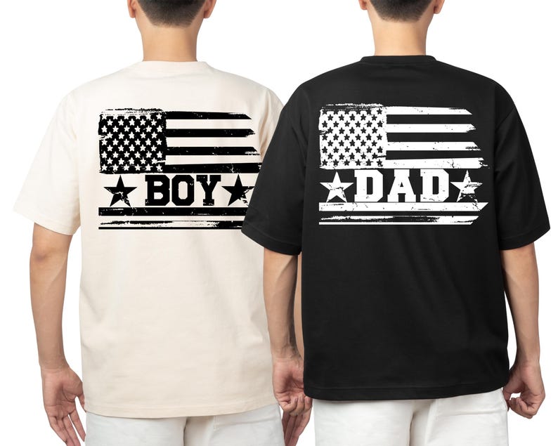 Puede incluir: Dos camisetas, una crema y otra negra, presentan un dise&ntilde;o de bandera americana desgastada en la espalda. La camiseta crema dice "BOY" y la negra dice "DAD" en letras blancas. Ambas son de manga corta.
