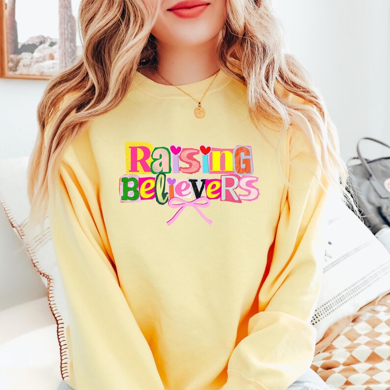 Peut inclure: Un sweat-shirt jaune clair avec l'inscription "Raising Believers" en lettres color&eacute;es. Le texte comprend un n&oelig;ud rose et des c&oelig;urs. Le sweat-shirt est de style d&eacute;contract&eacute; et confortable.