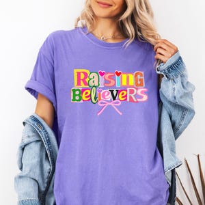 Raising Believers graphic shirt featuring playful layered typography with mixed font styles and decorative heart accents; the design displays the phrase Raising Believers in bold stacked lettering with textured details and a bow graphic centered beneath the text; the layout is balanced and easy to read with a cheerful faith inspired aesthetic; the artwork is centered on the shirt creating a clean modern graphic suitable for everyday wear