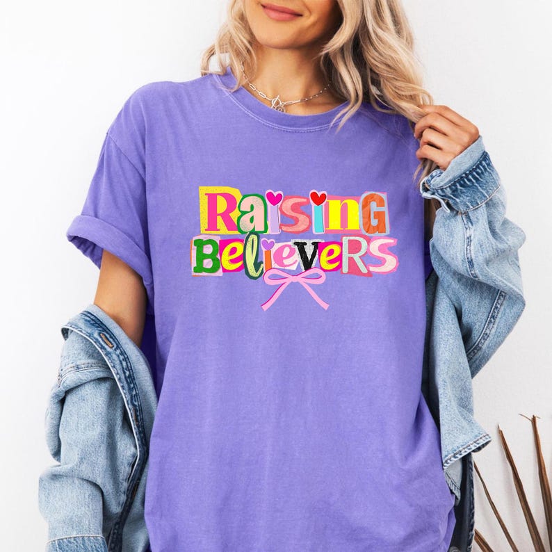 Raising Believers graphic shirt featuring playful layered typography with mixed font styles and decorative heart accents; the design displays the phrase Raising Believers in bold stacked lettering with textured details and a bow graphic centered beneath the text; the layout is balanced and easy to read with a cheerful faith inspired aesthetic; the artwork is centered on the shirt creating a clean modern graphic suitable for everyday wear