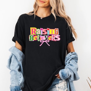 Peut inclure: T-shirt noir avec l'inscription "Raising Believers" en lettres multicolores. Le motif comprend des c&oelig;urs et un n&oelig;ud rose. Le t-shirt est de style d&eacute;contract&eacute;.