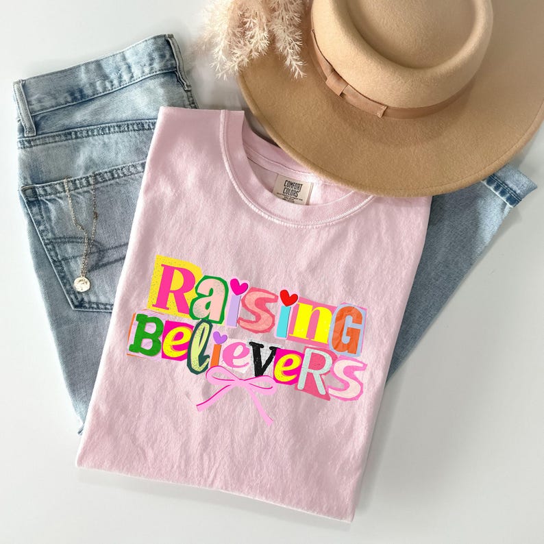 Raising Believers graphic shirt featuring playful layered typography with mixed font styles and decorative heart accents; the design displays the phrase Raising Believers in bold stacked lettering with textured details and a bow graphic centered beneath the text; the layout is balanced and easy to read with a cheerful faith inspired aesthetic; the artwork is centered on the shirt creating a clean modern graphic suitable for everyday wear
