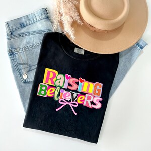 Peut inclure: Un t-shirt noir avec le texte "Raising Believers" en lettres color&eacute;es, un n&oelig;ud rose et deux c&oelig;urs. Le t-shirt est pos&eacute; sur un jean bleu clair, un chapeau beige et une brindille de fleurs s&eacute;ch&eacute;es.