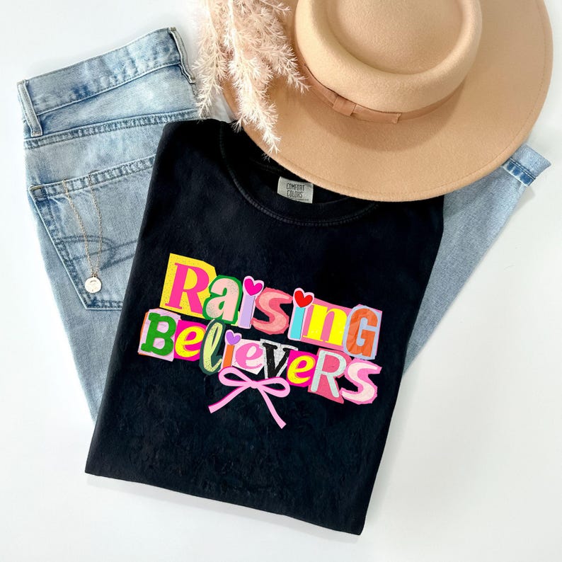 Peut inclure: Un t-shirt noir avec le texte "Raising Believers" en lettres color&eacute;es, un n&oelig;ud rose et deux c&oelig;urs. Le t-shirt est pos&eacute; sur un jean bleu clair, un chapeau beige et une brindille de fleurs s&eacute;ch&eacute;es.