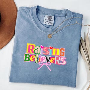 Raising Believers graphic shirt featuring playful layered typography with mixed font styles and decorative heart accents; the design displays the phrase Raising Believers in bold stacked lettering with textured details and a bow graphic centered beneath the text; the layout is balanced and easy to read with a cheerful faith inspired aesthetic; the artwork is centered on the shirt creating a clean modern graphic suitable for everyday wear