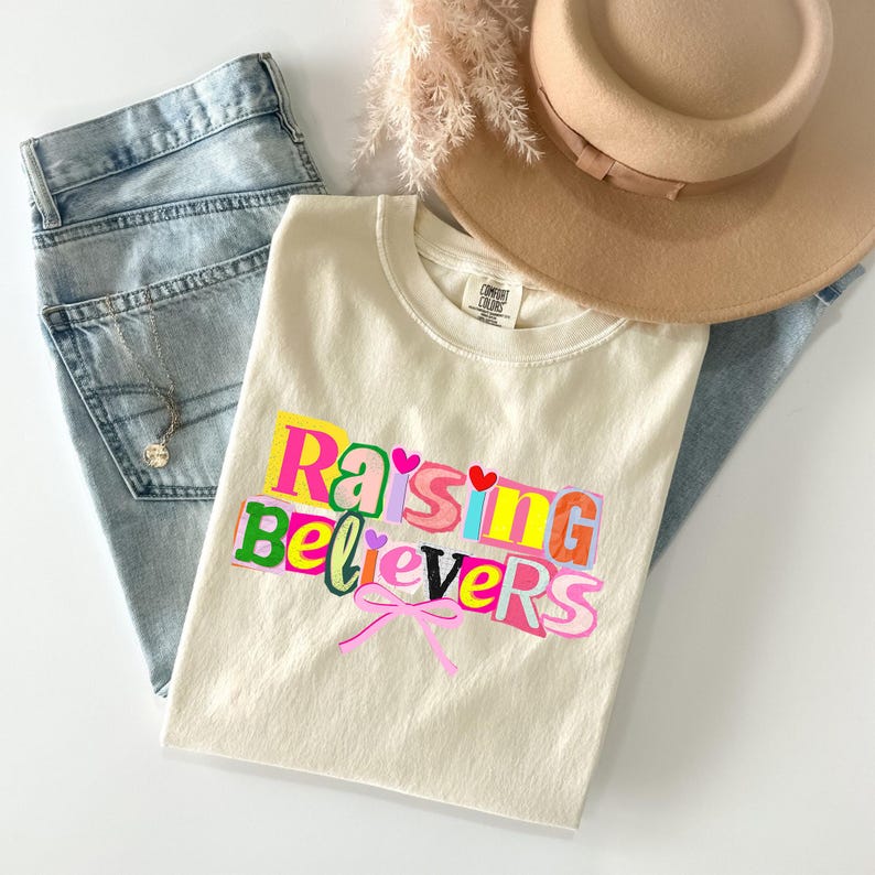 Peut inclure: T-shirt cr&egrave;me avec l'inscription multicolore "Raising Believers". Il est associ&eacute; &agrave; un jean en denim d&eacute;lav&eacute;, un chapeau beige et un collier d&eacute;licat. Un n&oelig;ud rose est &eacute;galement pr&eacute;sent.