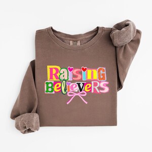 Peut inclure: Un sweat-shirt marron avec l'inscription "Raising Believers" en lettres color&eacute;es. Les lettres sont bord&eacute;es de diff&eacute;rentes couleurs, avec des c&oelig;urs et des n&oelig;uds. Les manches sont retrouss&eacute;es et le sweat-shirt est pli&eacute;.