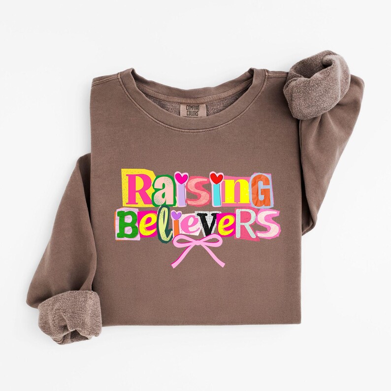 Peut inclure: Un sweat-shirt marron avec l'inscription "Raising Believers" en lettres color&eacute;es. Les lettres sont bord&eacute;es de diff&eacute;rentes couleurs, avec des c&oelig;urs et des n&oelig;uds. Les manches sont retrouss&eacute;es et le sweat-shirt est pli&eacute;.