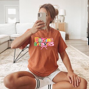 Raising Believers graphic shirt featuring playful layered typography with mixed font styles and decorative heart accents; the design displays the phrase Raising Believers in bold stacked lettering with textured details and a bow graphic centered beneath the text; the layout is balanced and easy to read with a cheerful faith inspired aesthetic; the artwork is centered on the shirt creating a clean modern graphic suitable for everyday wear
