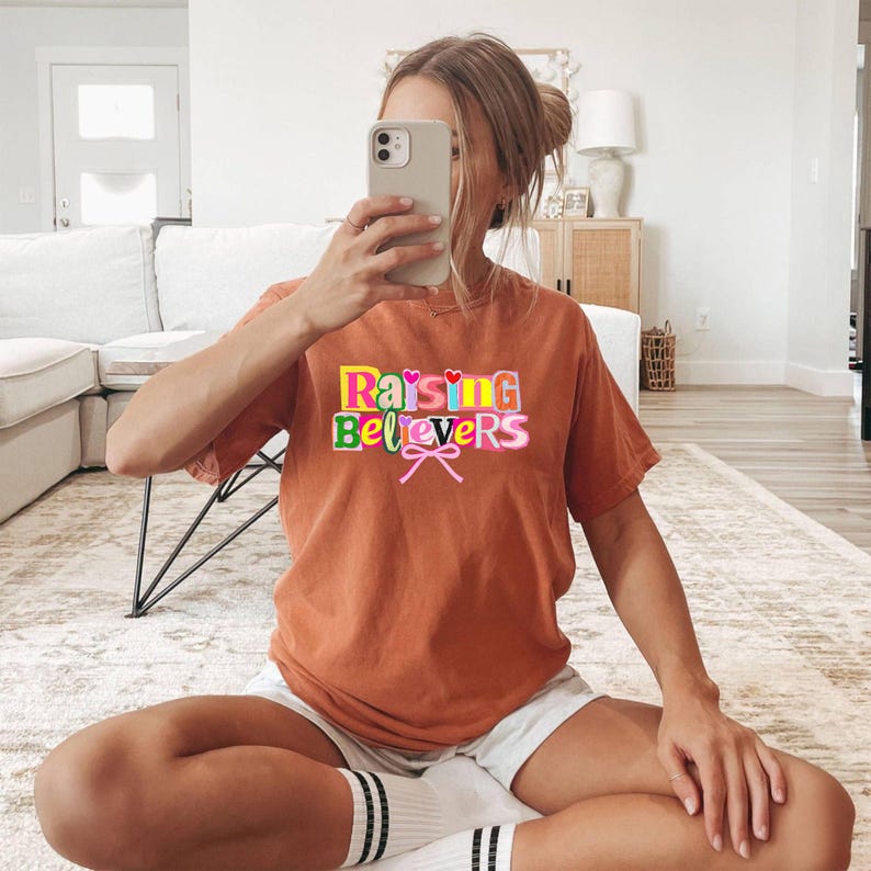 Raising Believers graphic shirt featuring playful layered typography with mixed font styles and decorative heart accents; the design displays the phrase Raising Believers in bold stacked lettering with textured details and a bow graphic centered beneath the text; the layout is balanced and easy to read with a cheerful faith inspired aesthetic; the artwork is centered on the shirt creating a clean modern graphic suitable for everyday wear