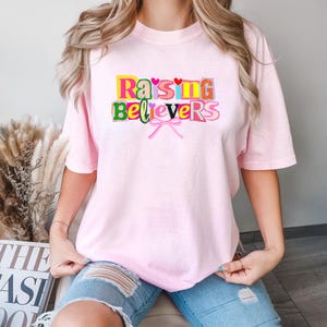 Peut inclure: Un t-shirt rose clair avec l'inscription "Raising Believers" en lettres color&eacute;es. Le motif comprend un n&oelig;ud rose et de petits c&oelig;urs. Le t-shirt est de style d&eacute;contract&eacute;, &agrave; manches courtes.