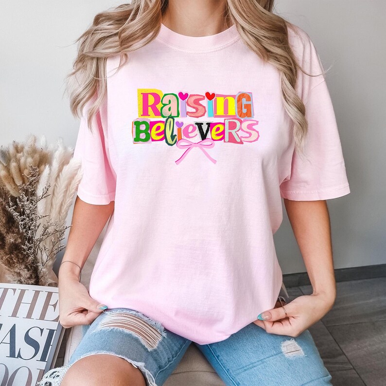 Peut inclure: Un t-shirt rose clair avec l'inscription "Raising Believers" en lettres color&eacute;es. Le motif comprend un n&oelig;ud rose et de petits c&oelig;urs. Le t-shirt est de style d&eacute;contract&eacute;, &agrave; manches courtes.