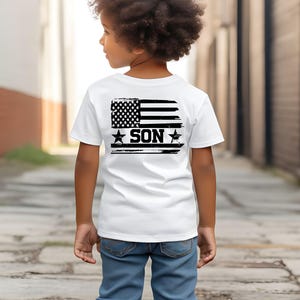 Puede incluir: Una camiseta blanca con un dise&ntilde;o de bandera estadounidense en blanco y negro desgastado en la espalda, con la palabra "SON" en letras negritas. La camiseta la lleva un ni&ntilde;o, combinada con vaqueros azules.