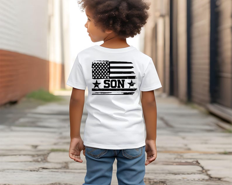 Puede incluir: Una camiseta blanca con un dise&ntilde;o de bandera estadounidense en blanco y negro desgastado en la espalda, con la palabra "SON" en letras negritas. La camiseta la lleva un ni&ntilde;o, combinada con vaqueros azules.