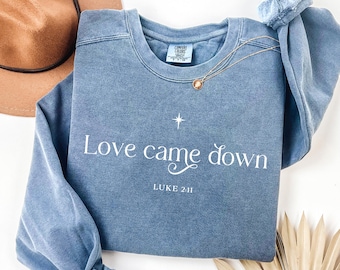 May include: A blue crewneck sweatshirt with the words "Love came down" and "Luke 2:11" printed in white. A gold necklace and a scrunchie are on the sweatshirt. A brown hat is in the background.