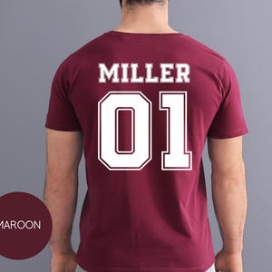 May include: Maroon t-shirt with the name "MILLER" and the number "01" in white, outlined lettering on the back. The shirt is a solid maroon color, and the text is in a bold, collegiate style font. The word "MAROON" is in a maroon circle.