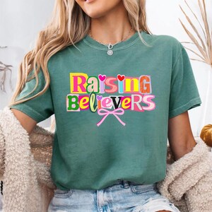 Raising Believers graphic shirt featuring playful layered typography with mixed font styles and decorative heart accents; the design displays the phrase Raising Believers in bold stacked lettering with textured details and a bow graphic centered beneath the text; the layout is balanced and easy to read with a cheerful faith inspired aesthetic; the artwork is centered on the shirt creating a clean modern graphic suitable for everyday wear