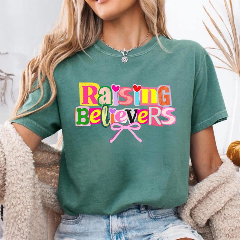 Raising Believers graphic shirt featuring playful layered typography with mixed font styles and decorative heart accents; the design displays the phrase Raising Believers in bold stacked lettering with textured details and a bow graphic centered beneath the text; the layout is balanced and easy to read with a cheerful faith inspired aesthetic; the artwork is centered on the shirt creating a clean modern graphic suitable for everyday wear